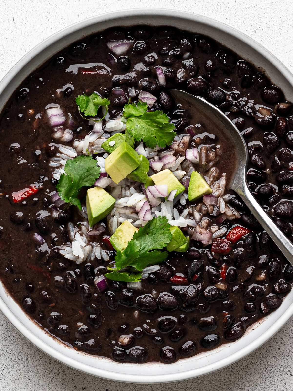 Closeup of Instant Pot Cuban Black Bean soup served in a white bowl garnished with diced red onion, diced avocado, and white rice