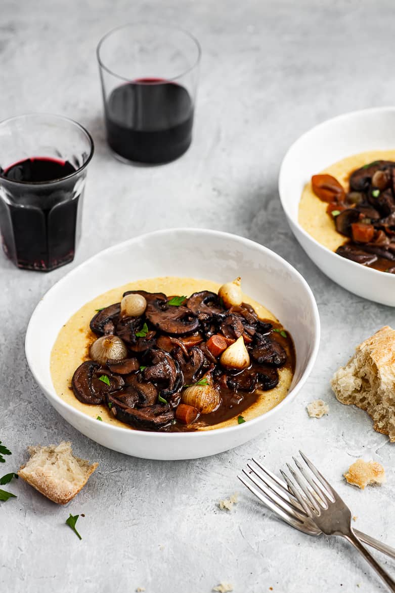 Mushroom Bourguignon in bowl served over creamy polenta with crusty bread and wine glasses