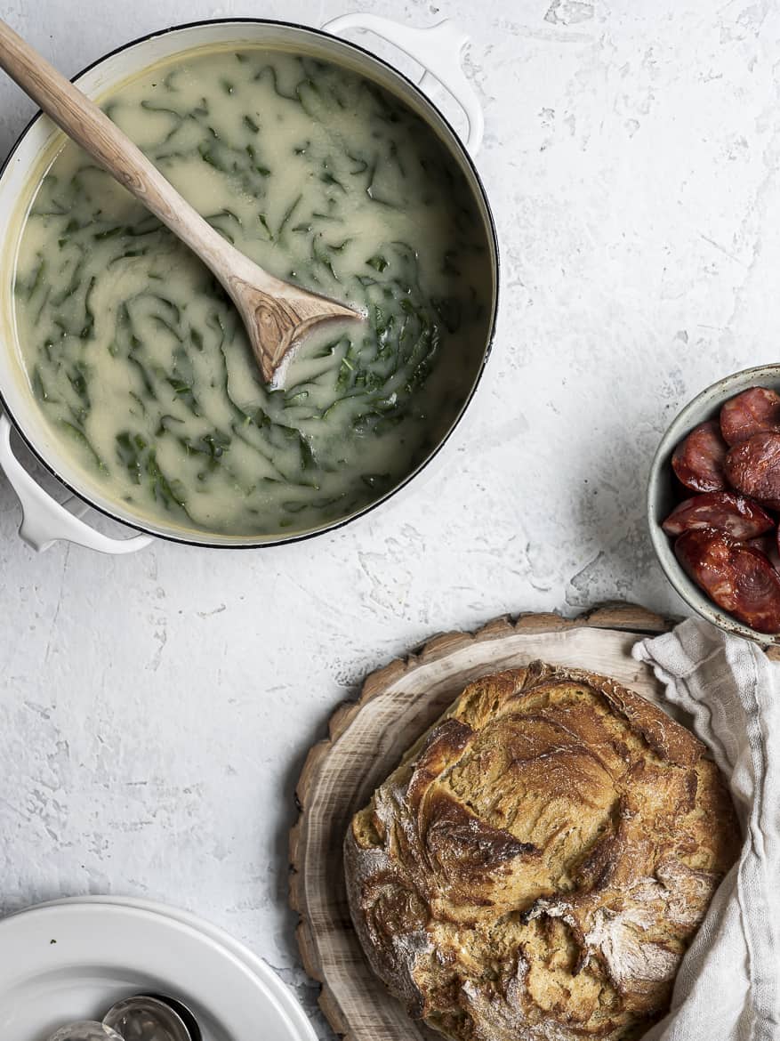 Caldo verde in soup pot shown with a Portuguese bread loaf and choriço sausage in bowl