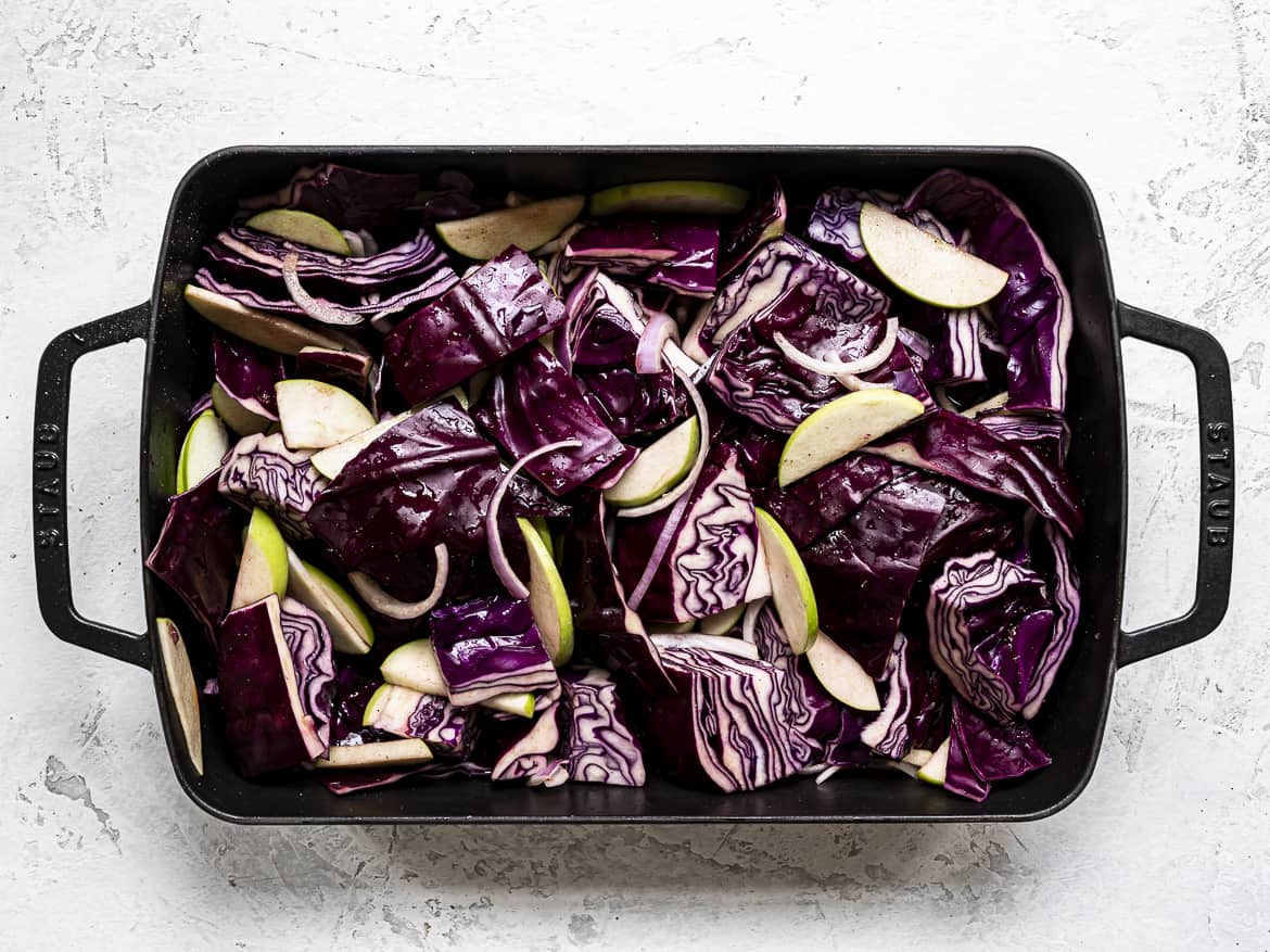 Chopped red cabbage in roasting pan with sliced apples, onions, mixed with apple cider and balsamic vinegar