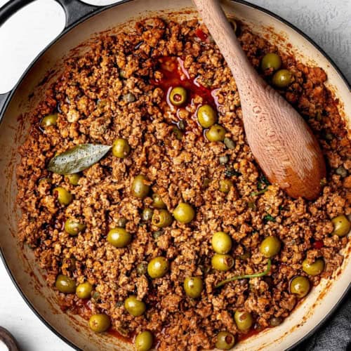 Finished dish of turkey picadillo with olives in saucepan and a wooden spoon for serving.
