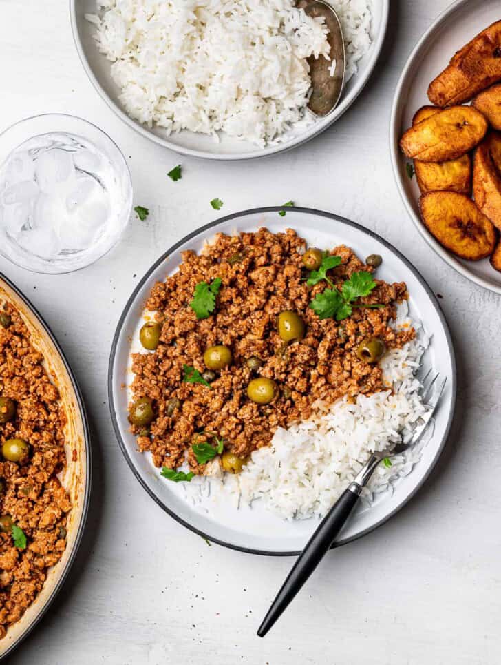 Turkey picadillo served on a white plate with bowls of plantains and white rice on the side. 