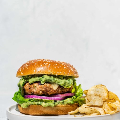 One poblano turkey burger filled with lettuce, red onion slices, and avocado sauce on a white plate with chips on the side.