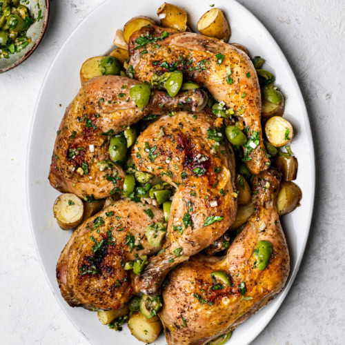 Chicken legs, leeks, and potatoes on a platter topped with green olive salsa