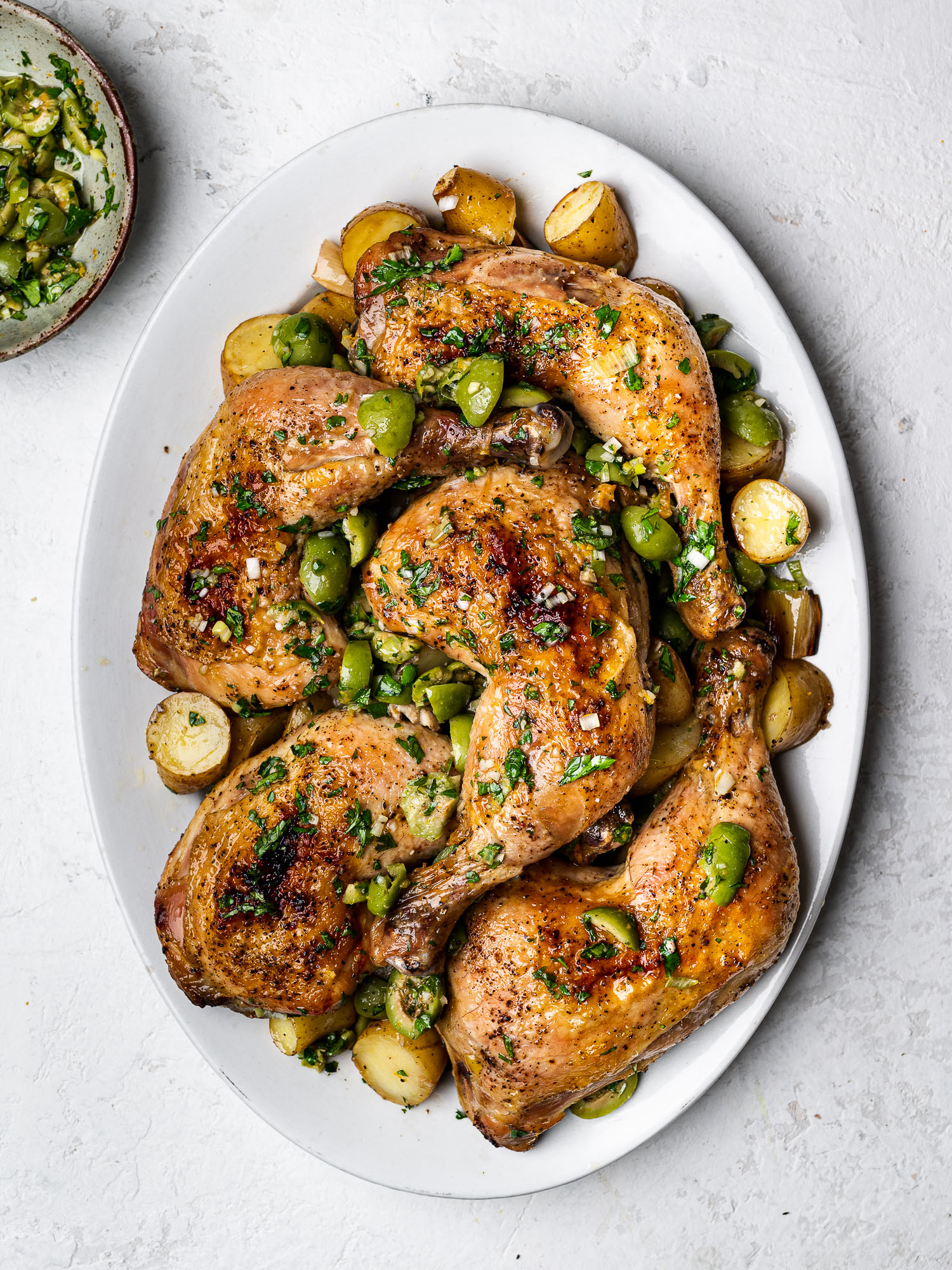 Chicken legs, leeks, and potatoes on a platter topped with green olive salsa 