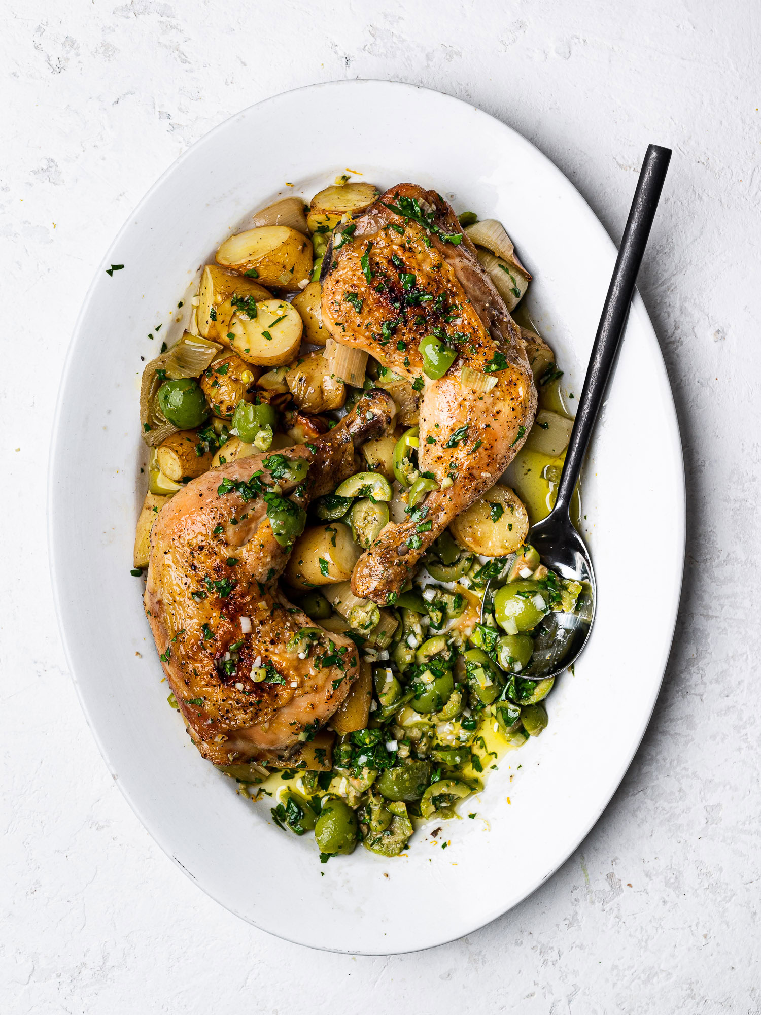 Chicken legs, leeks, and potatoes on a platter topped with green olive salsa