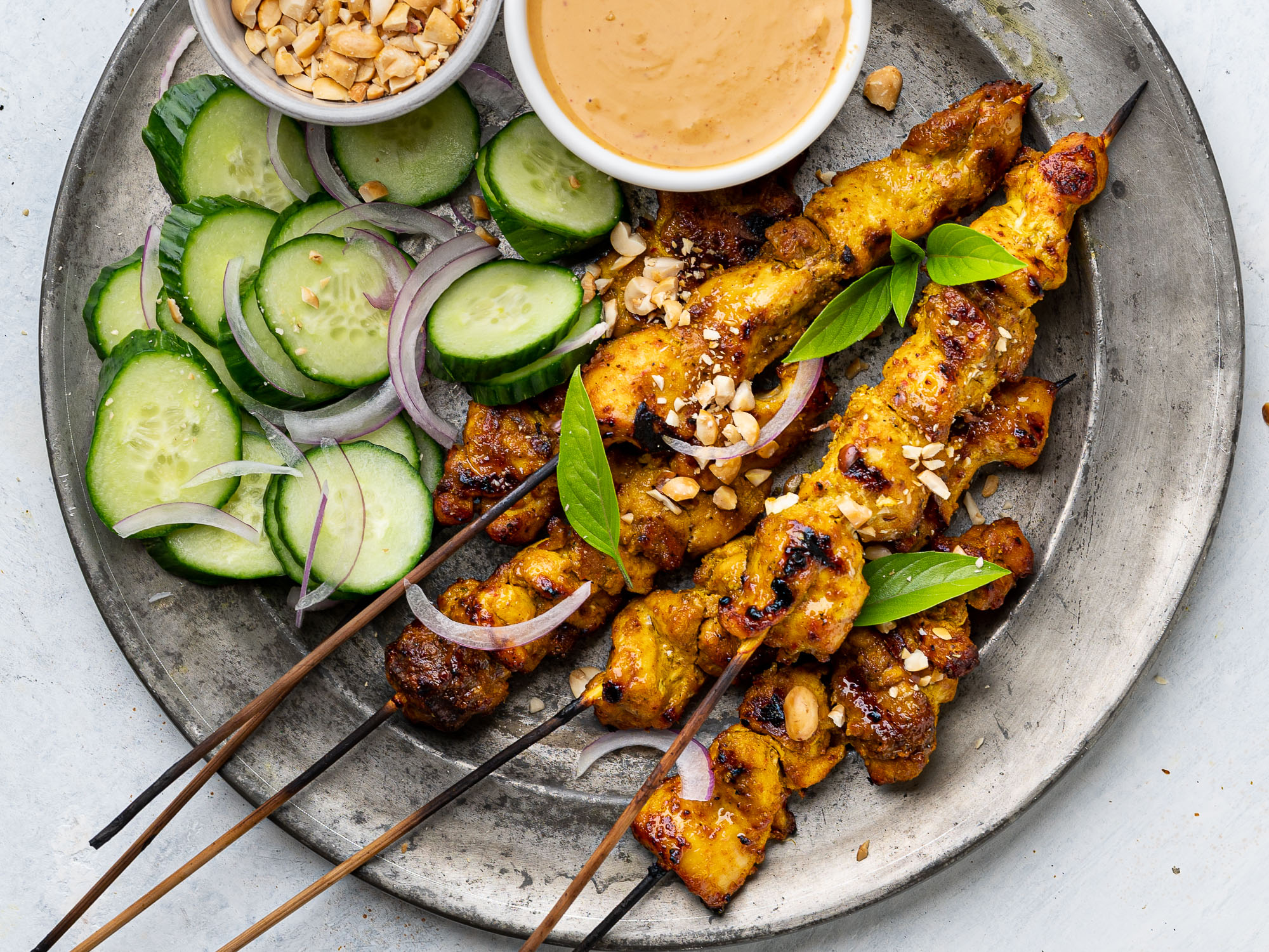 close up of chicken satay skewers on a plate with peanut dipping sauce and cucumber slices