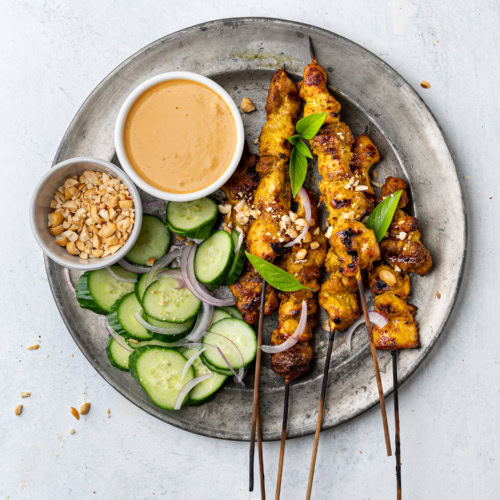 chicken satay skewers on a plate with peanut dipping sauce and cucumber slices