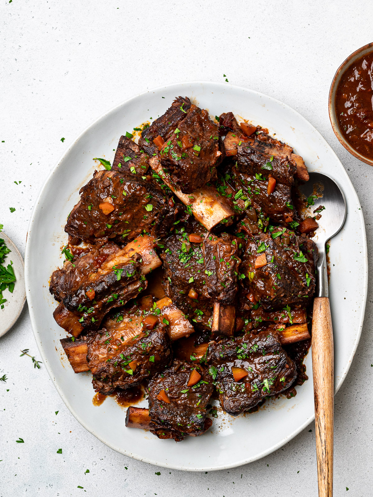 Braised short ribs served on a platter garnished with chopped parsley and sauce on the side in a bowl