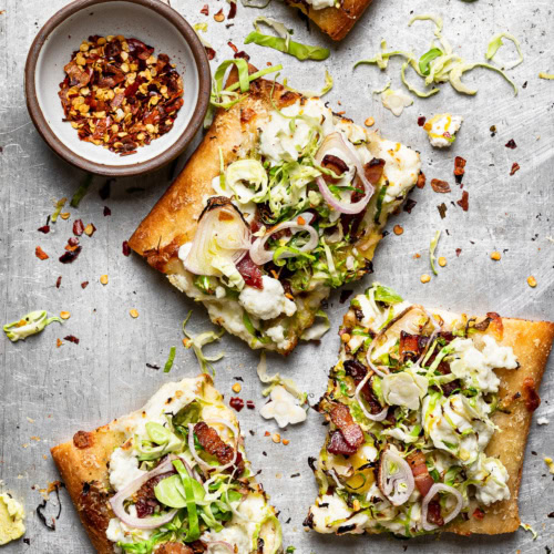 Brussels Sprouts and Ricotta Flatbread Pizza pieces on baking sheet with bowl of pepper flakes on the side