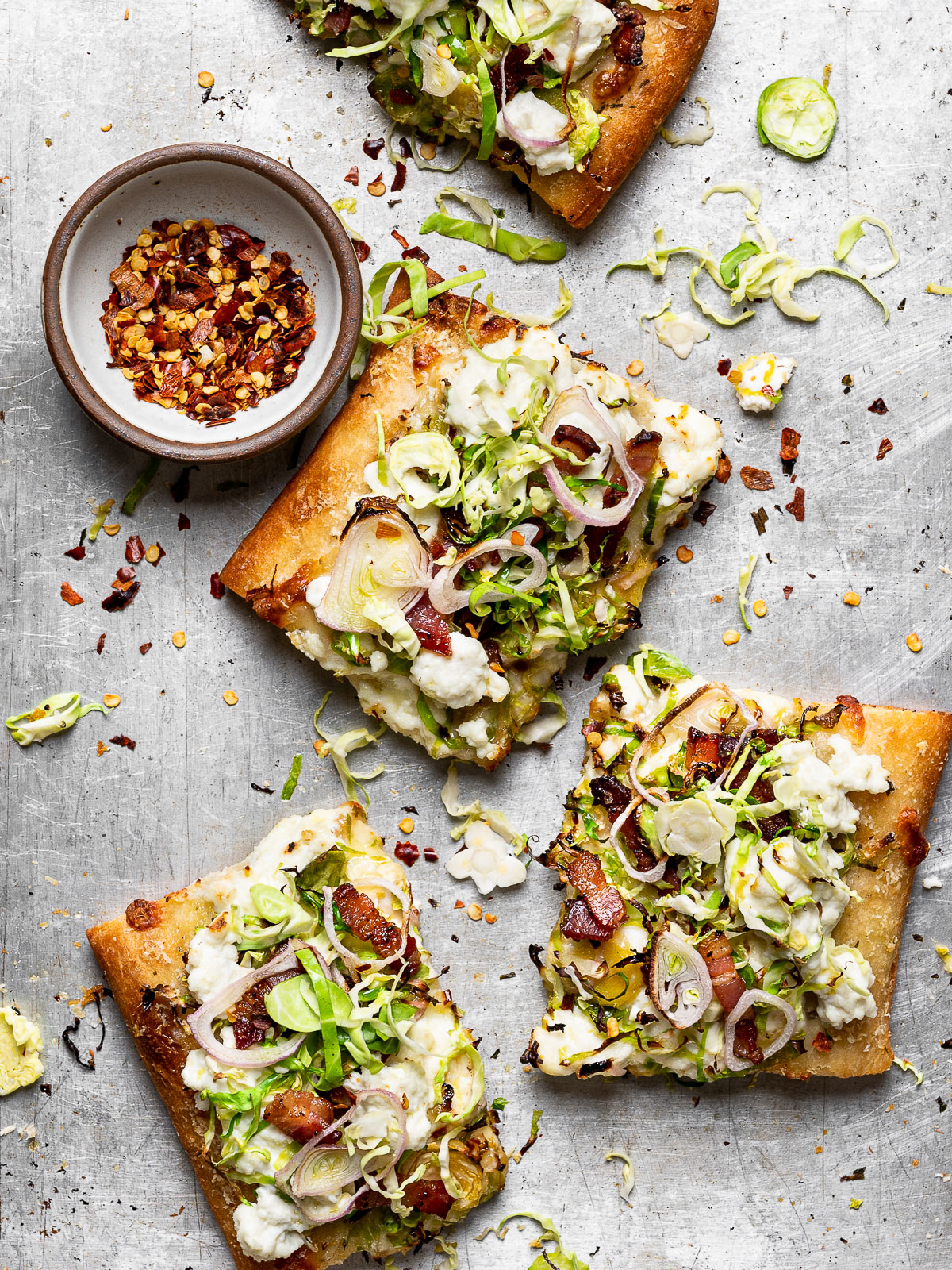 Brussels Sprouts and Ricotta Flatbread Pizza pieces on baking sheet with bowl of pepper flakes on the side