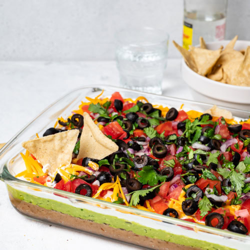 side view of 7 layer dip in glass dish with bowl of tortilla chips in the background