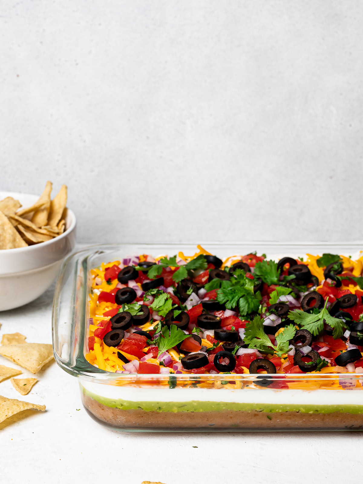 side view of 7 layer dip in glass dish with bowl of tortilla chips in the background