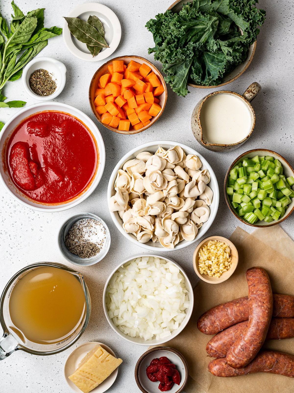 ingredients for recipe in bowls: sausage, onions, carrots, celery, garlic tortellini, broth, heavy cream, tomatoes and tomato paste, kale and spices