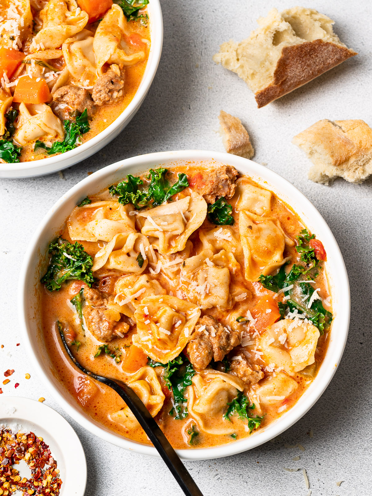 creamy tortellini soup with sausage and kale served in bowls and garnished with parmesan cheese and crushed red pepper flakes and crusty bread on the side