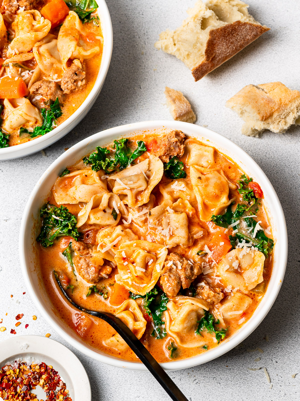 Creamy tortellini soup with sausage and kale served in bowls and garnished with parmesan cheese and crushed red pepper flakes and crusty bread on the side.