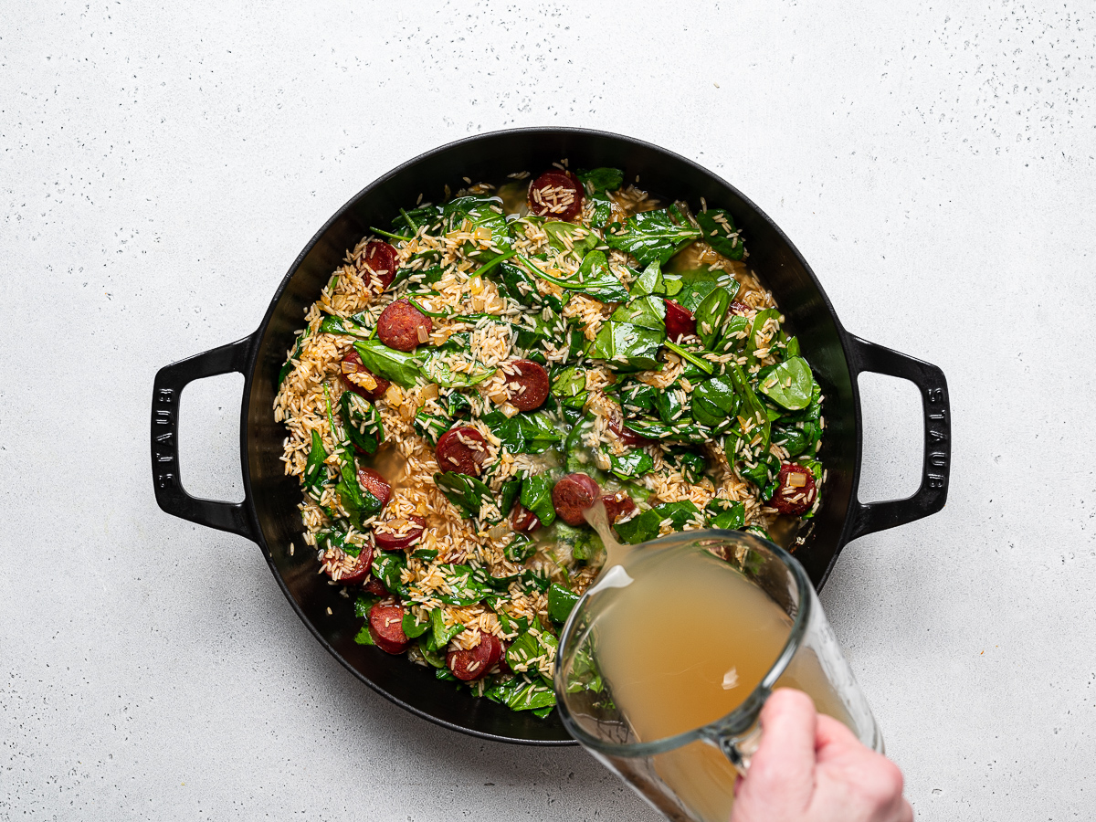 pouring broth into skillet with onions, garlic, rice, chorizo and spinach