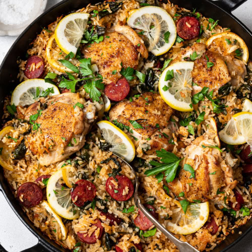 finished dish of chicken and chorizo rice dish in skillet with lemon slices and bowl of flakey salt on the side