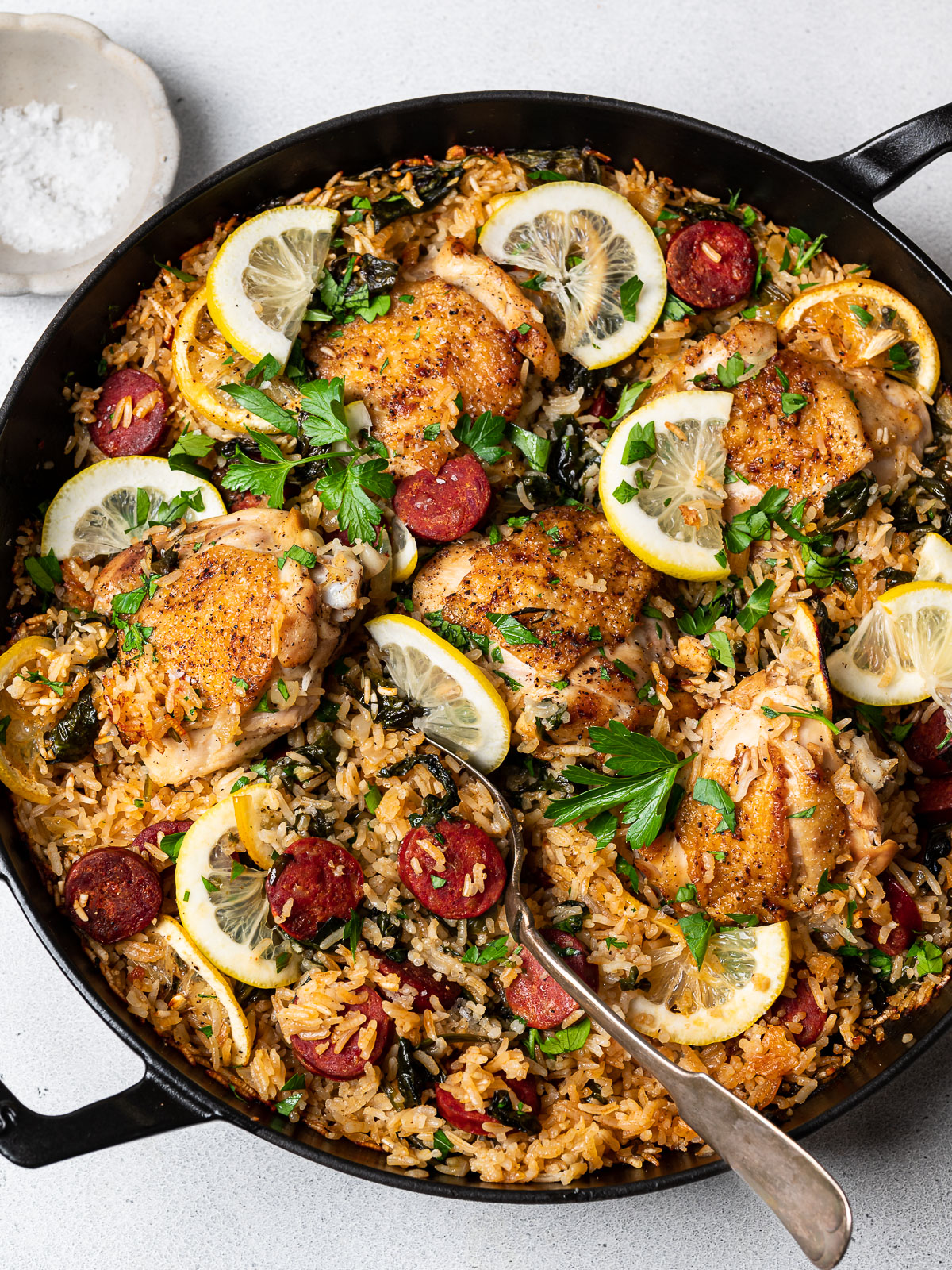 finished dish of chicken and chorizo rice dish in skillet with lemon slices and bowl of flakey salt on the side