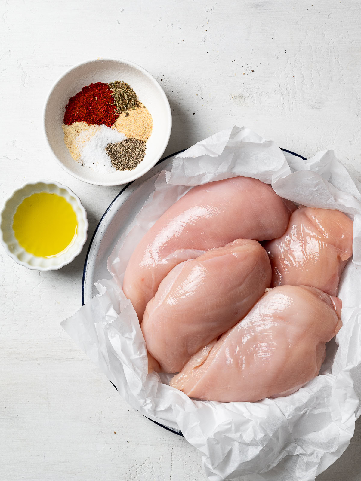 image of ingredients needed for recipe; chicken breasts on a plate, spices in a bowl, and olive oil in a bowl