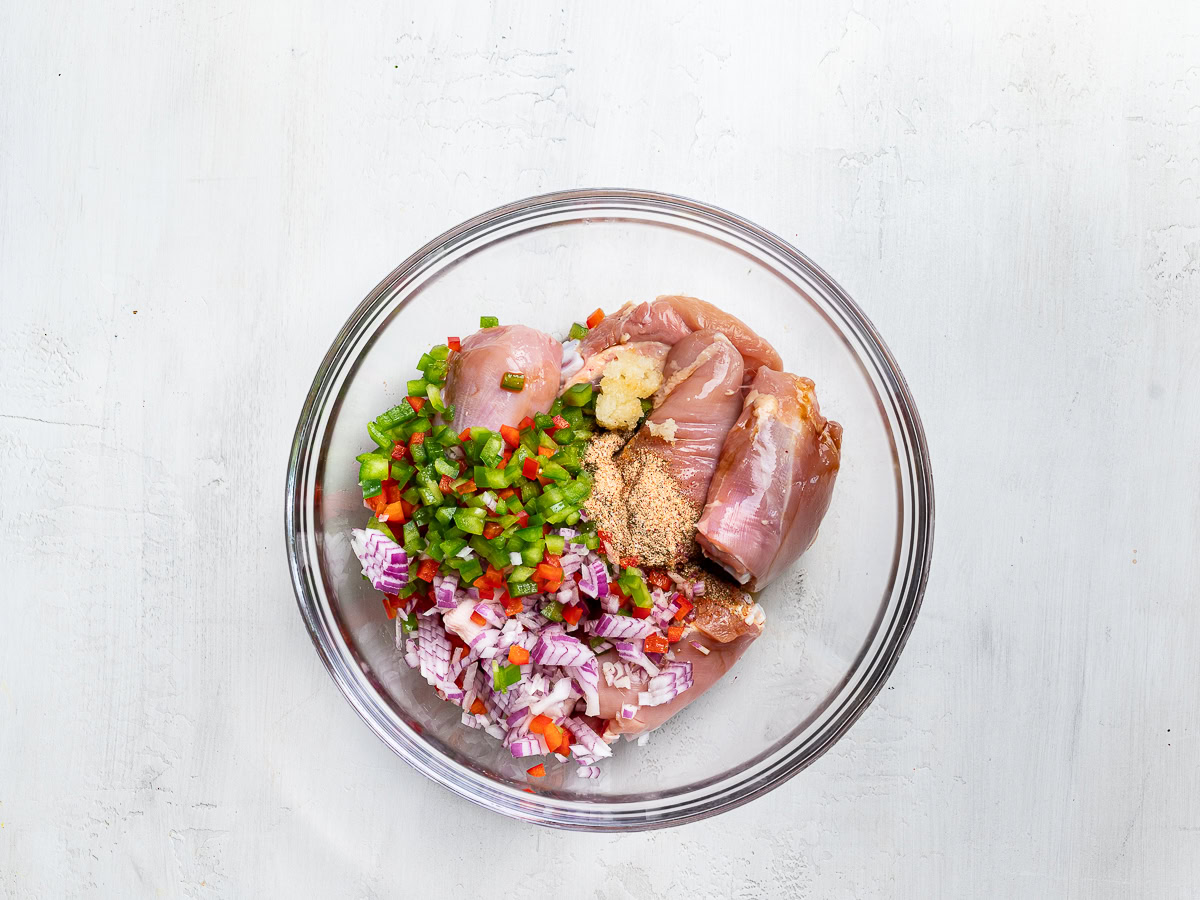 chicken pieces in glass mixing bowl with chopped onions, peppers, and garlic on top