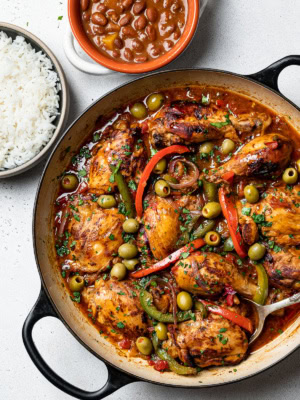 stewed chicken in skillet with tomato-based sauce with peppers, and olives and a bowl of beans and a bowl of rice on the side