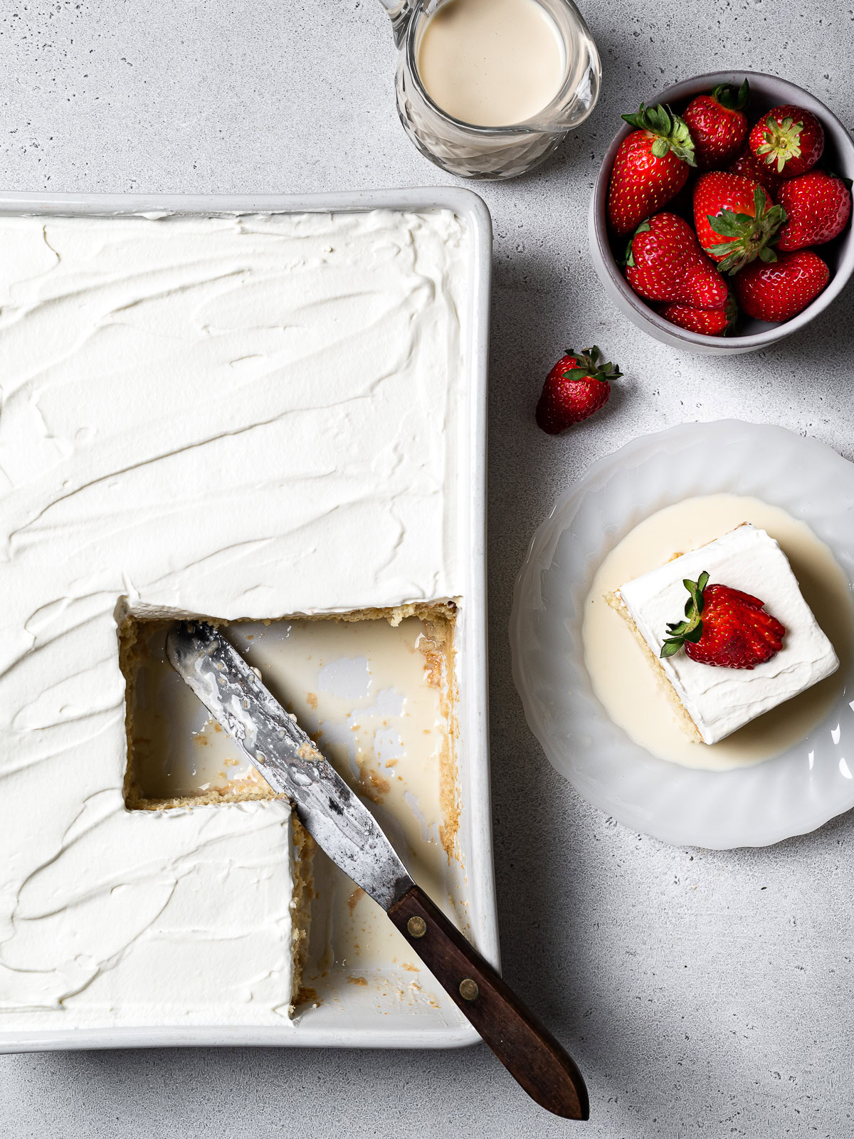 tres leches cake in baking dish with a slice on a plate, a bowl of strawberries, and a small pitcher with more milk syrup