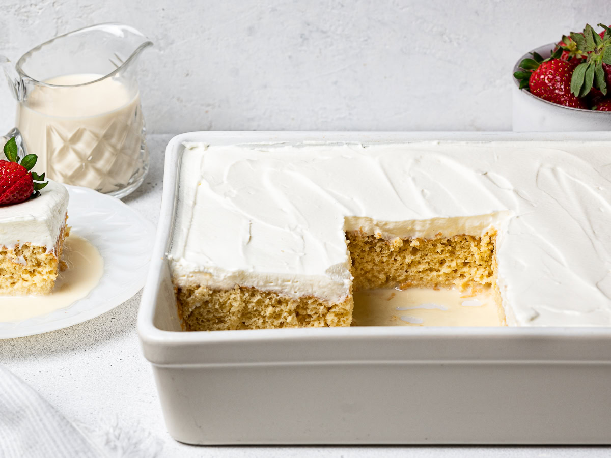 side-view of sliced tres leches cake in the baking dish with a small pitcher of milk syrup and bowl of strawberries on the side