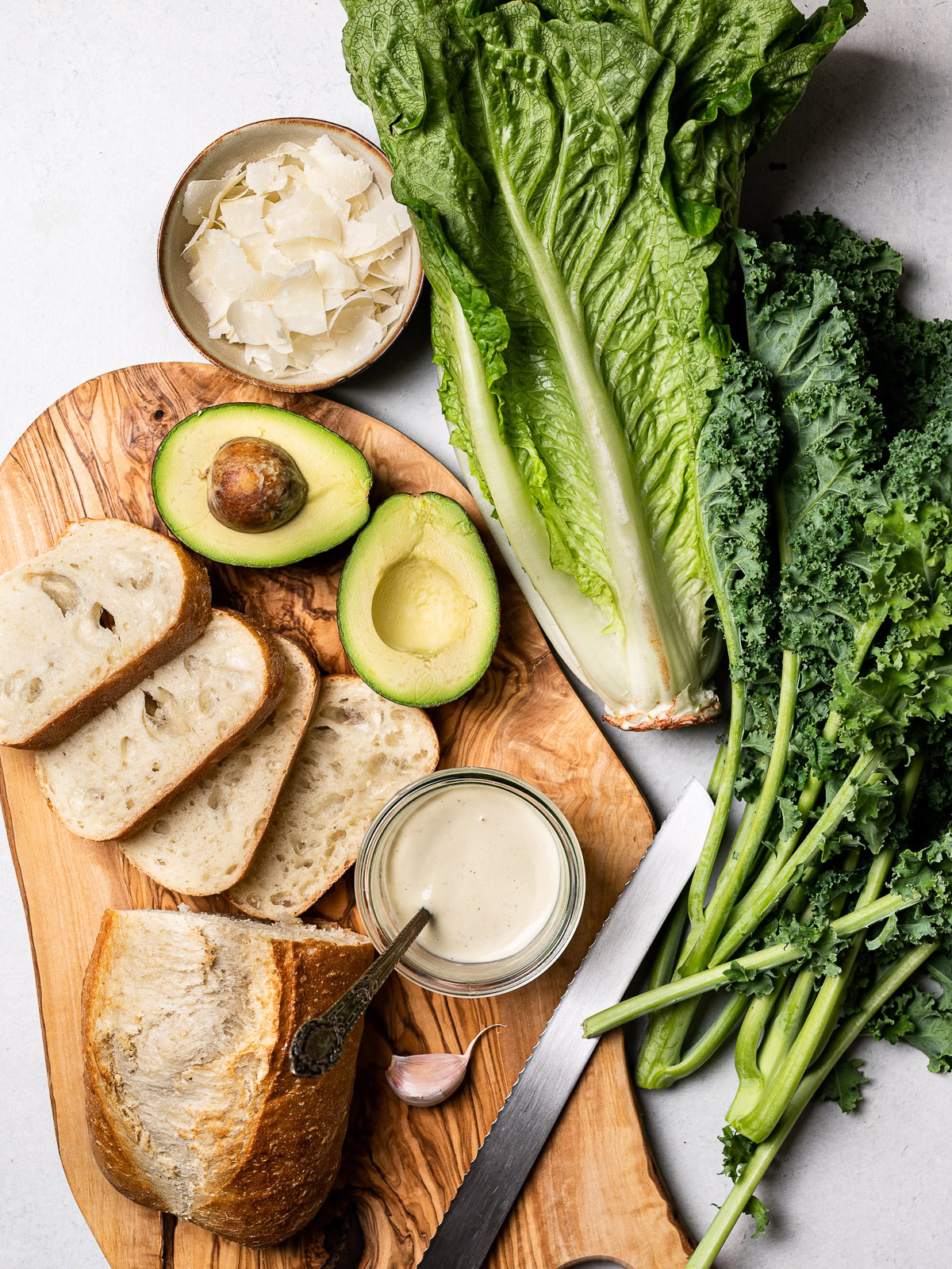 ingredients for kale Caesar salad; sliced bread, one bunch of kale, one romaine heart, 1 avocado, parmesan cheese curls in a bowl, and cashew Caesar dressing in a jar