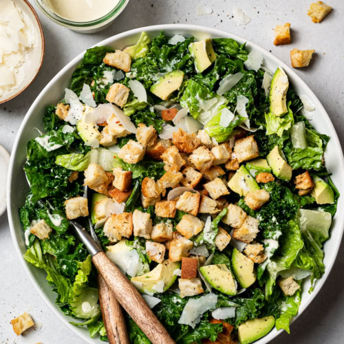 kale and romaine Caesar Salad in a bowl topped with croutons, avocado, and shaved parmesan