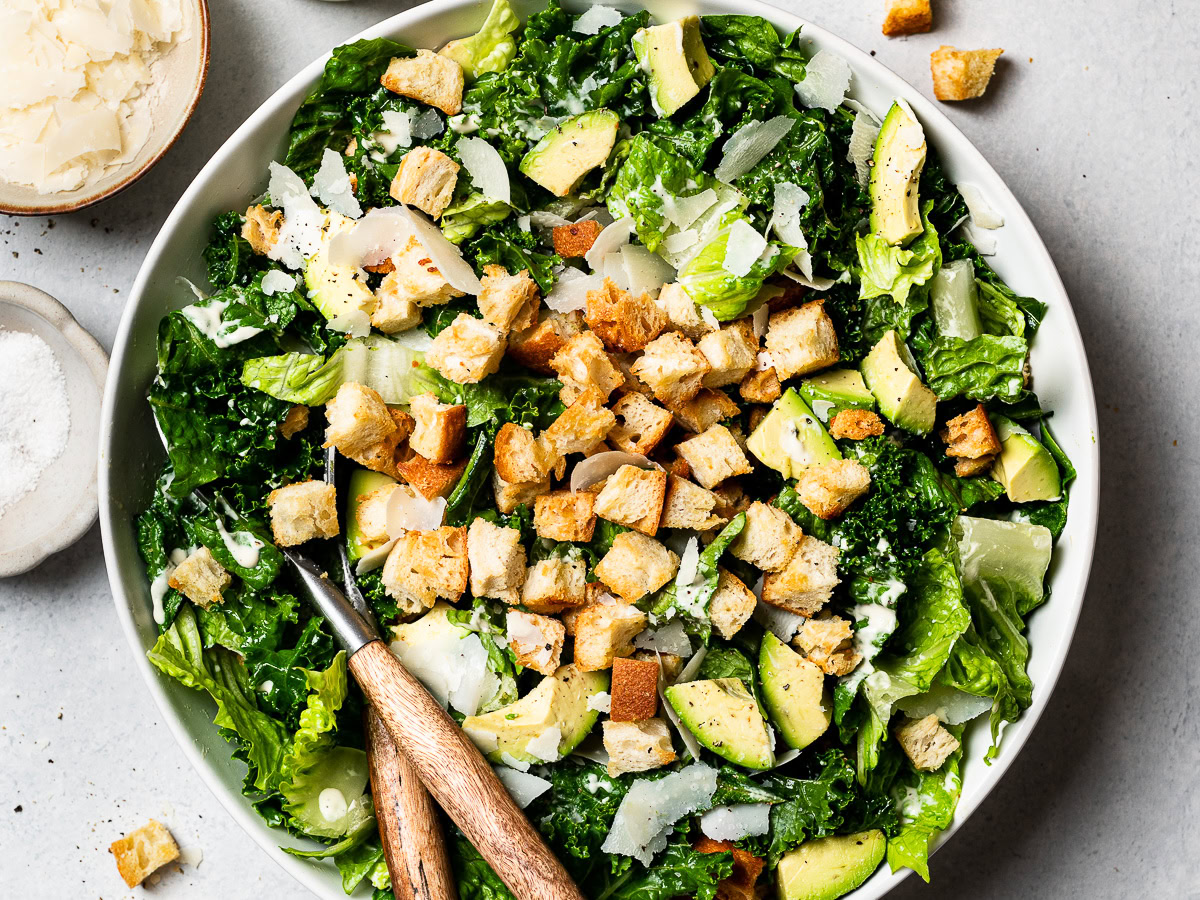 kale and romaine Caesar Salad in a bowl topped with croutons, avocado, and shaved parmesan