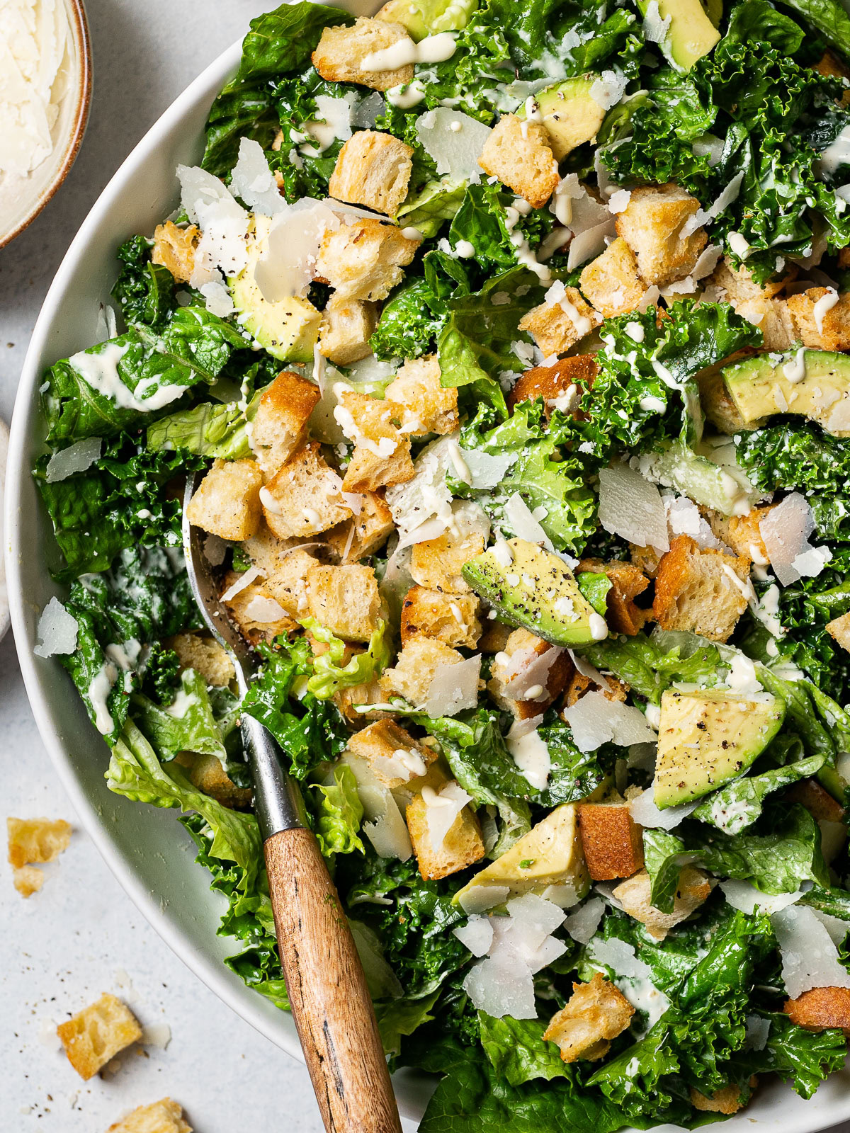 close up of kale and romaine Caesar Salad in a bowl topped with croutons, avocado, and shaved parmesan drizzled with more dressing