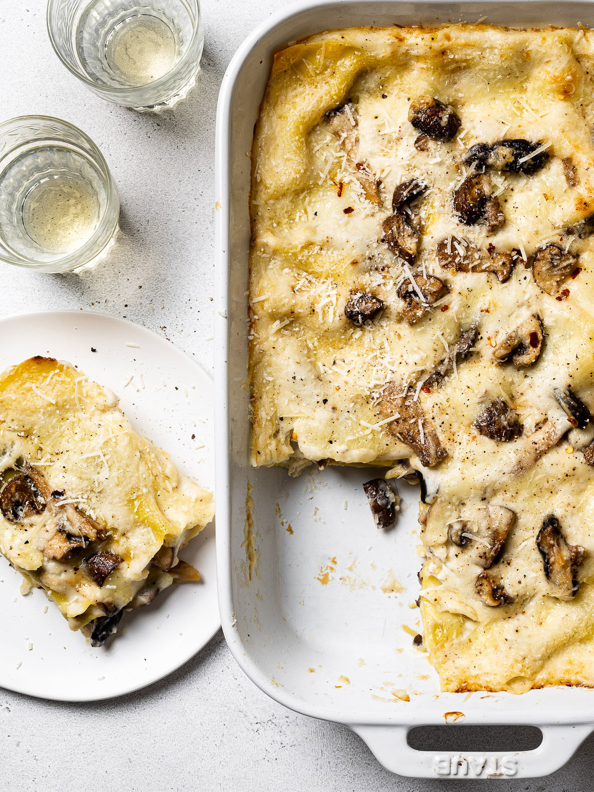 baked mushroom lasagna in baking dish with a served piece on the side and wine glasses