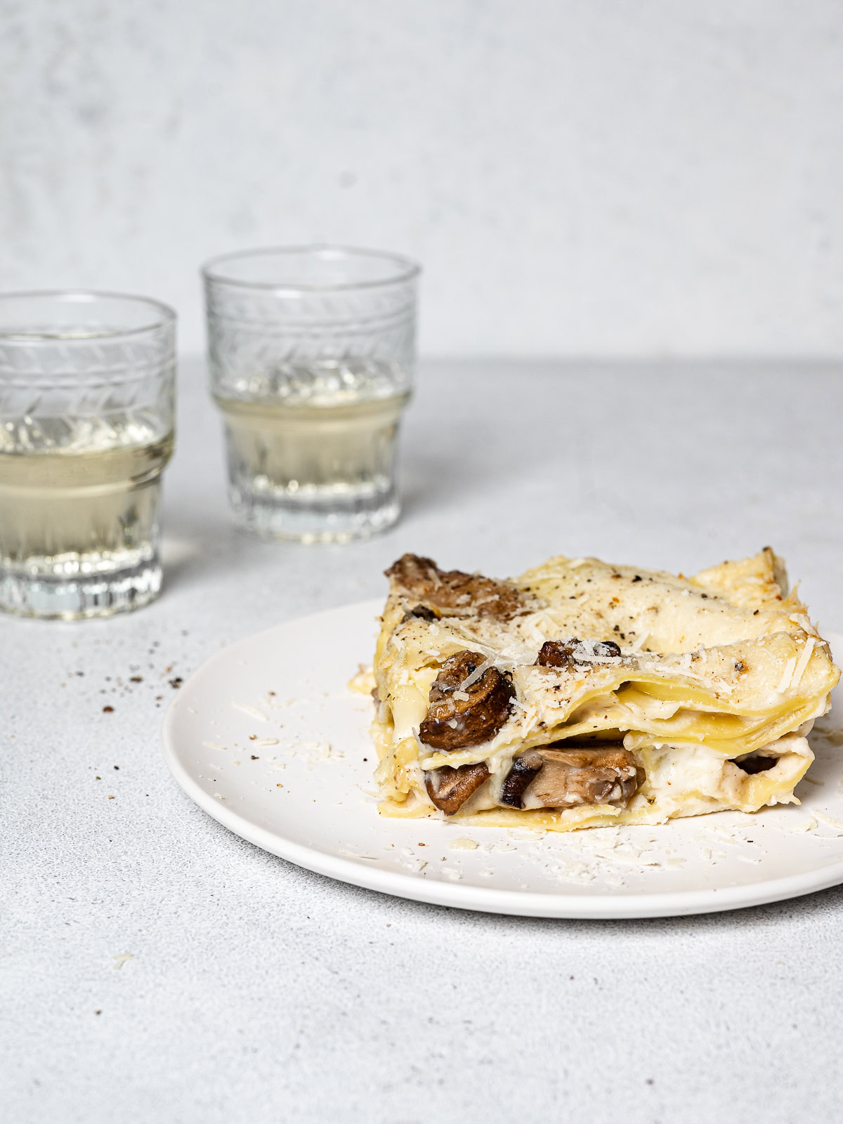 side view of mushroom lasagna serving on a plate with wine glasses in the background