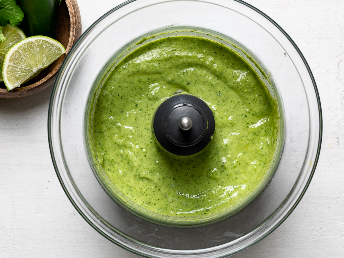 Food processor filled with blitzed green sauce. Jalapeños, limes, and cilantro are shown in a bowl on the side.
