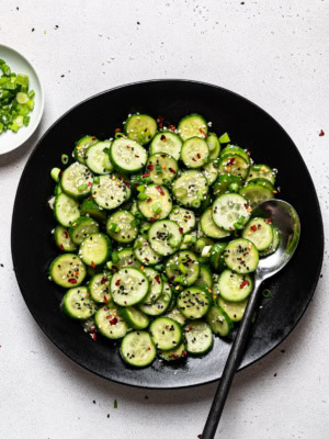 Sesame cucumber salad served in black bowl with chopped scallions on the side in a small bowl