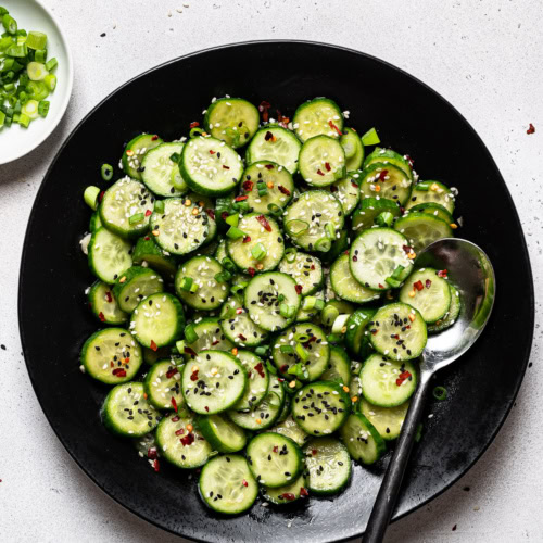 Sesame cucumber salad served in black bowl with chopped scallions on the side in a small bowl