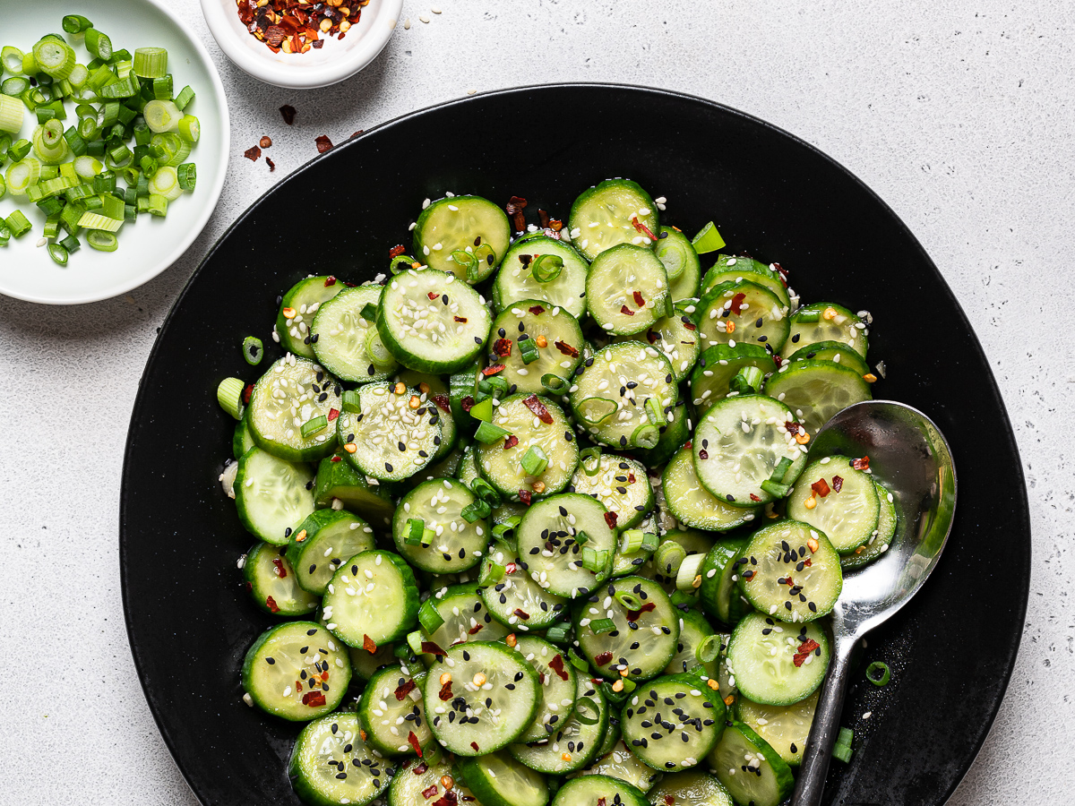 Sesame cucumber salad served in black bowl with chopped scallions and pepper flakes on the side in a small bowls