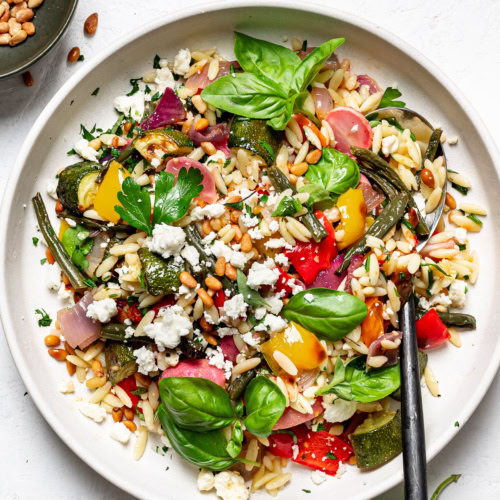 Orzo with roasted vegetables served on side dish with small bowl of pine nuts on the side
