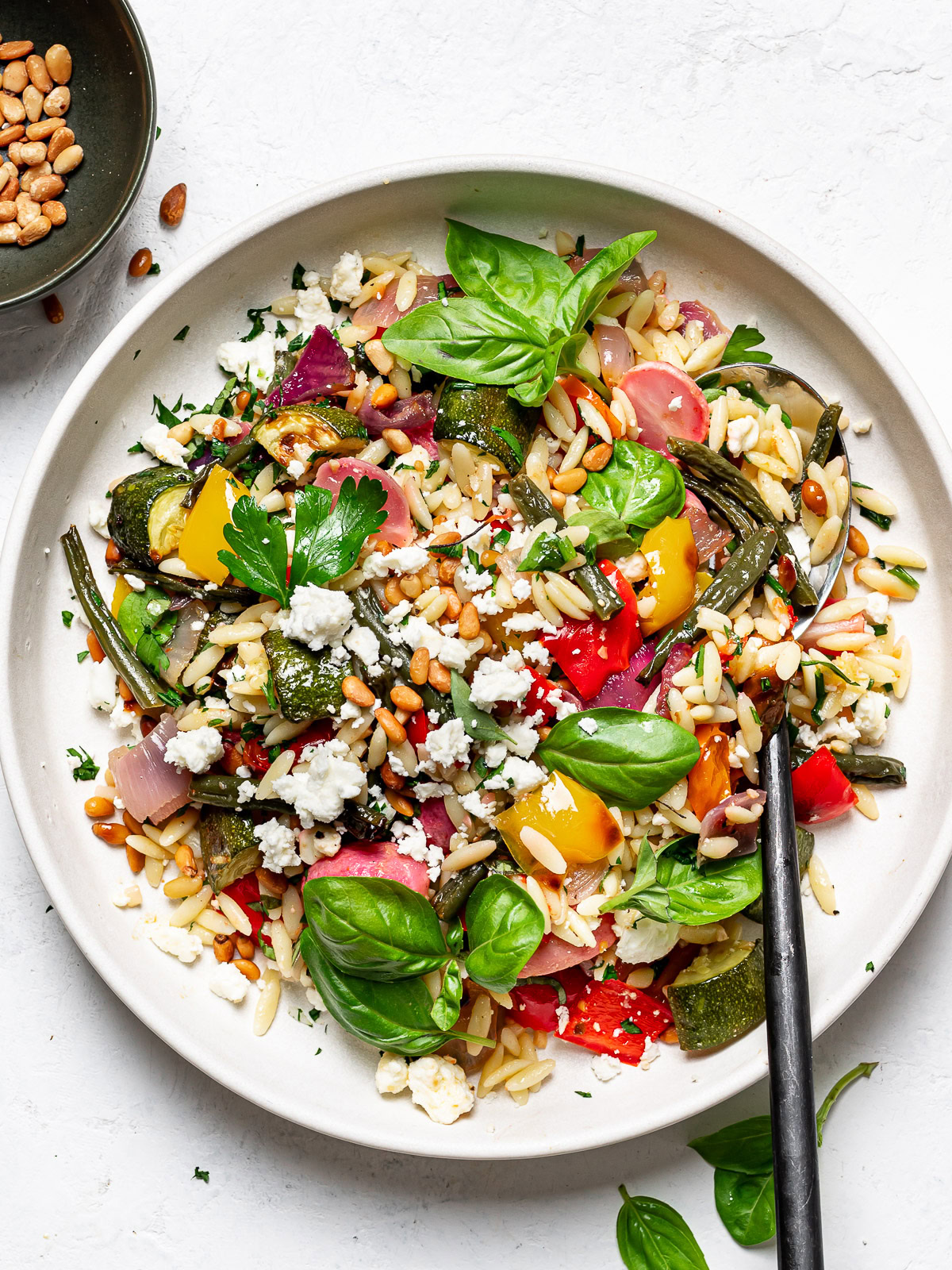 Orzo with roasted vegetables served on side dish with small bowl of pine nuts on the side