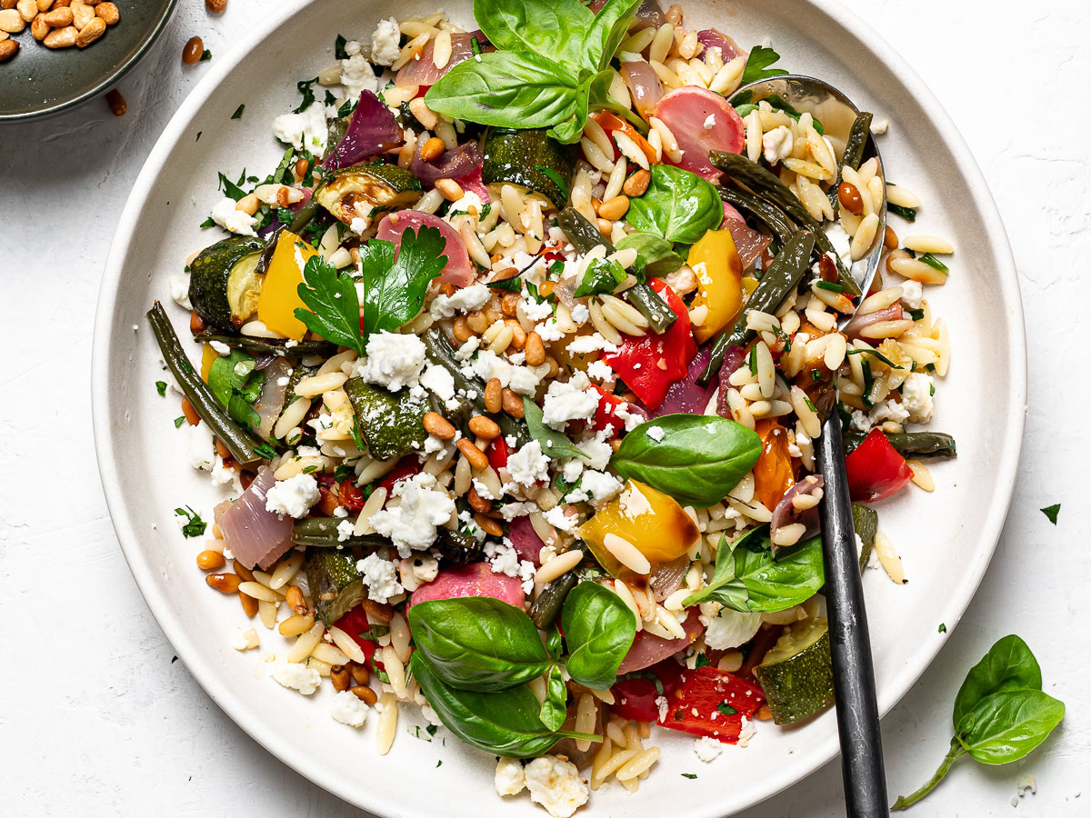 Orzo with roasted vegetables served on side dish with small bowl of pine nuts on the side