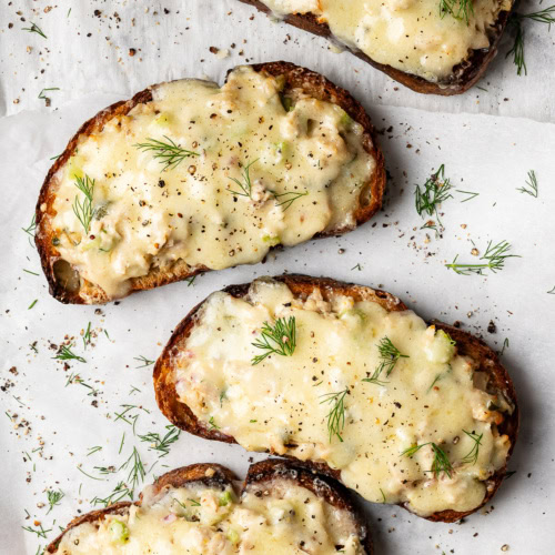 4 open-faced tuna melt toasts with melted cheese on baking sheet garnished with dill and black pepper