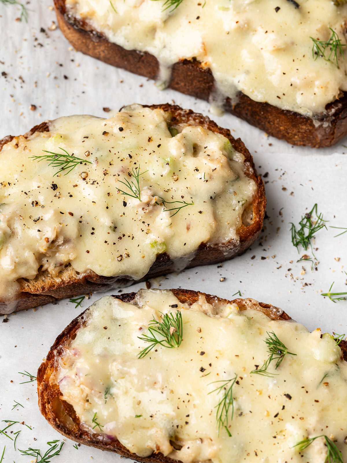 close up of open-faced tuna melt toasts with melted cheese on baking sheet garnished with dill and black pepper