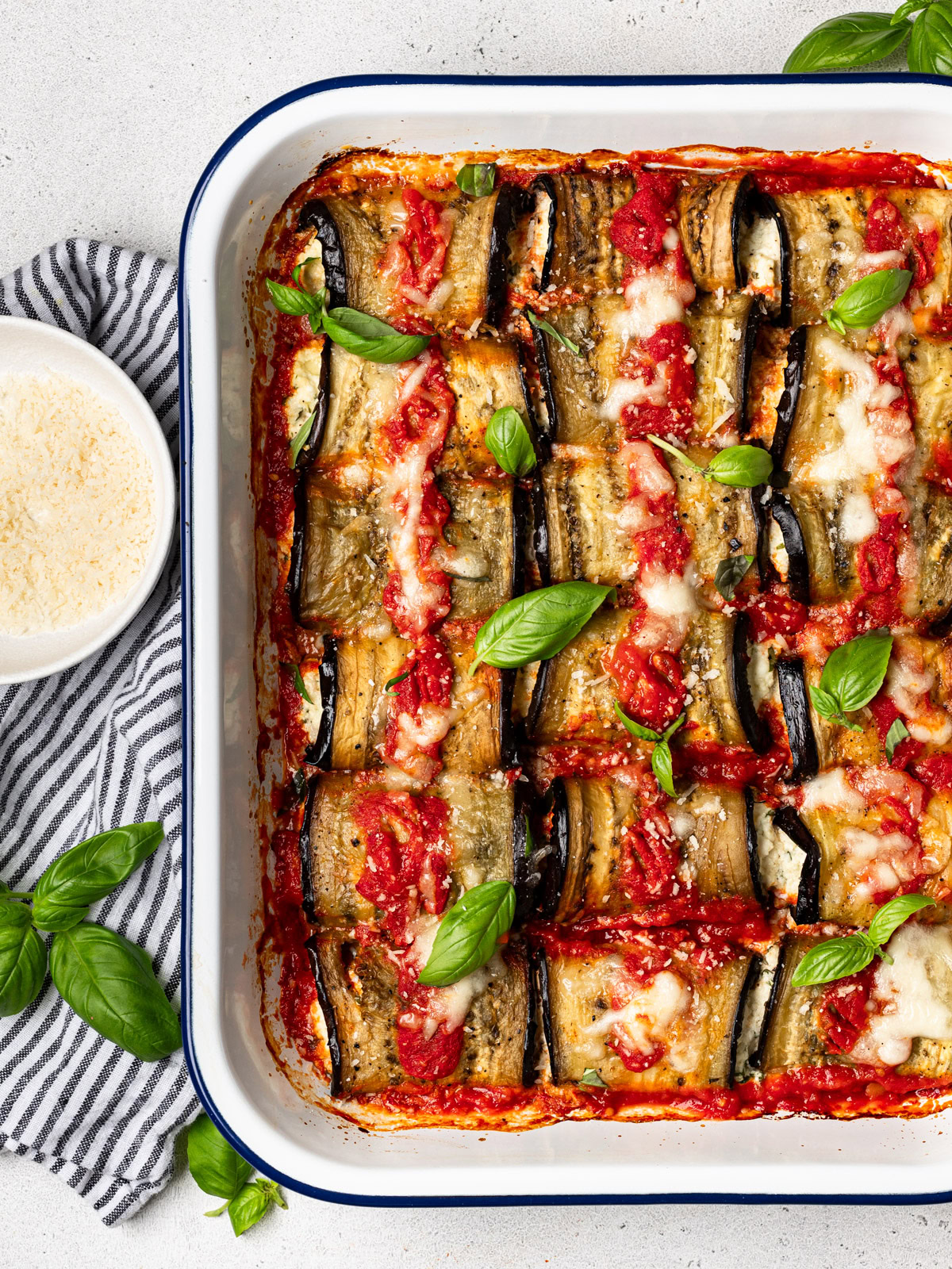 baked rollatini bundles  in casserole dish garnished with basil leaves and bowl of parmesan on the side