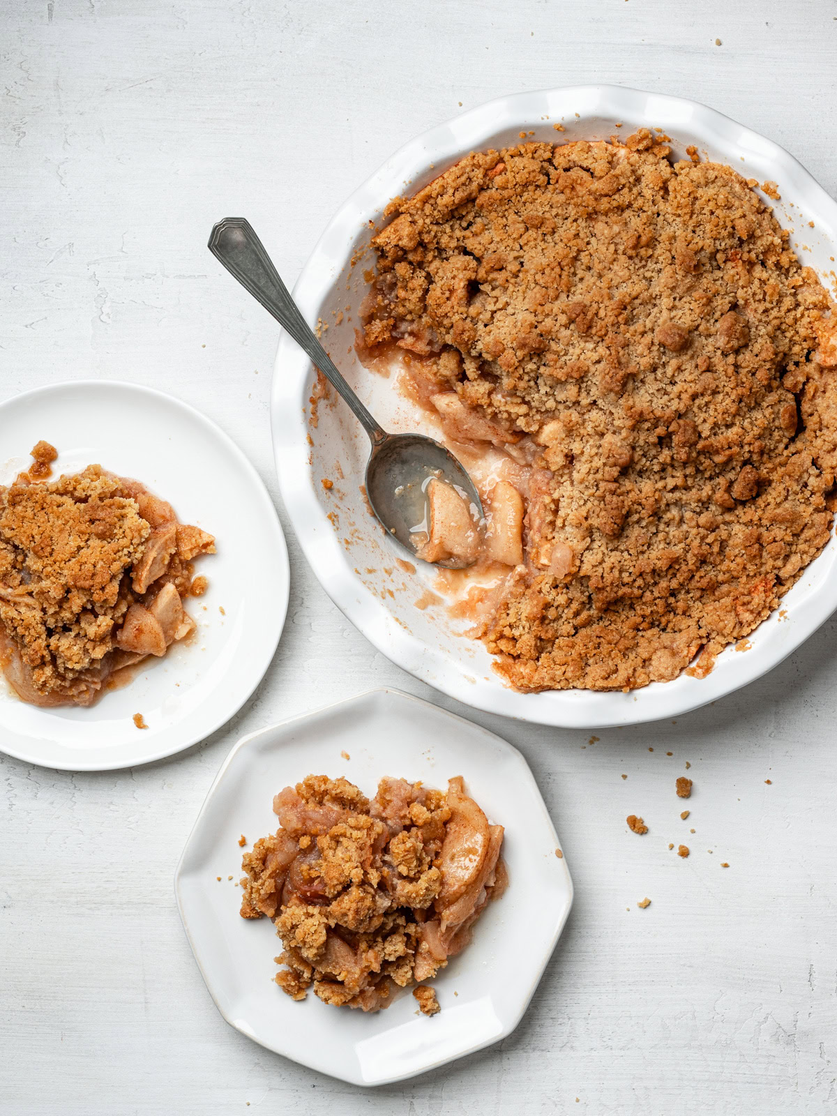 two servings of apple crisp without oats on small plates with pie in baking dish beside them