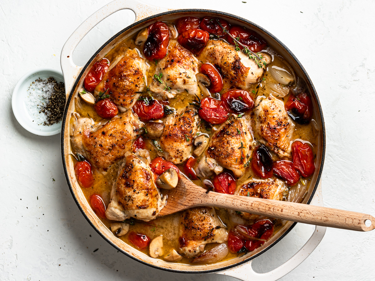 A finished dish of braised chicken with tomatoes and shallots in skillet. A wooden spoon is holding up a chicken piece.