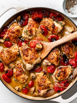 A finished dish of braised chicken with tomatoes and shallots in skillet. A wooden spoon is holding up a chicken piece.