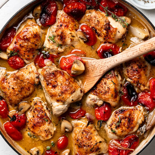 A finished dish of braised chicken with tomatoes and shallots in skillet. A wooden spoon is holding up a chicken piece.