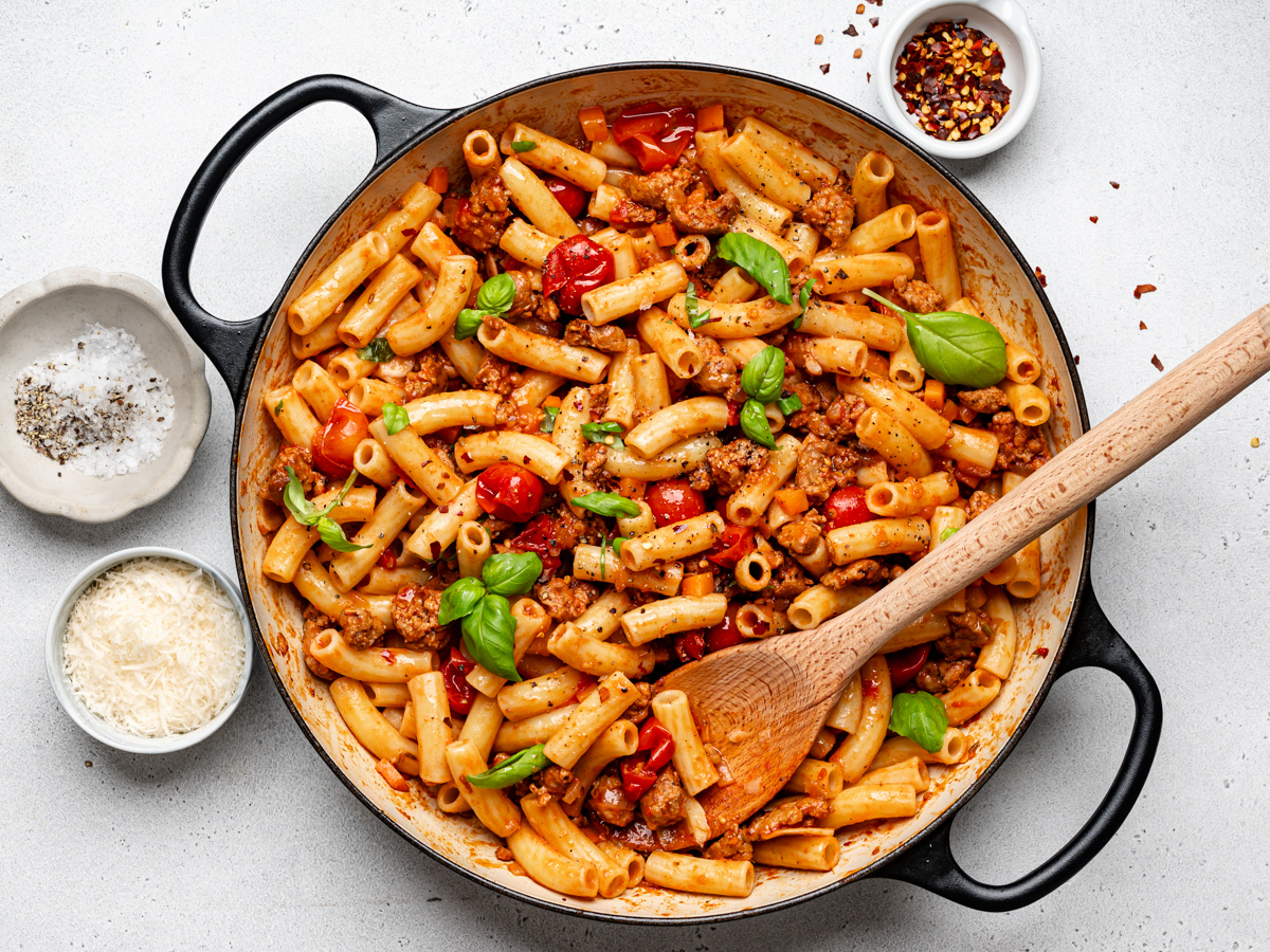 Spicy Italian Sausage pasta combined in skillet with small bowls of red pepper flakes, cheese and salt and pepper on the side. 