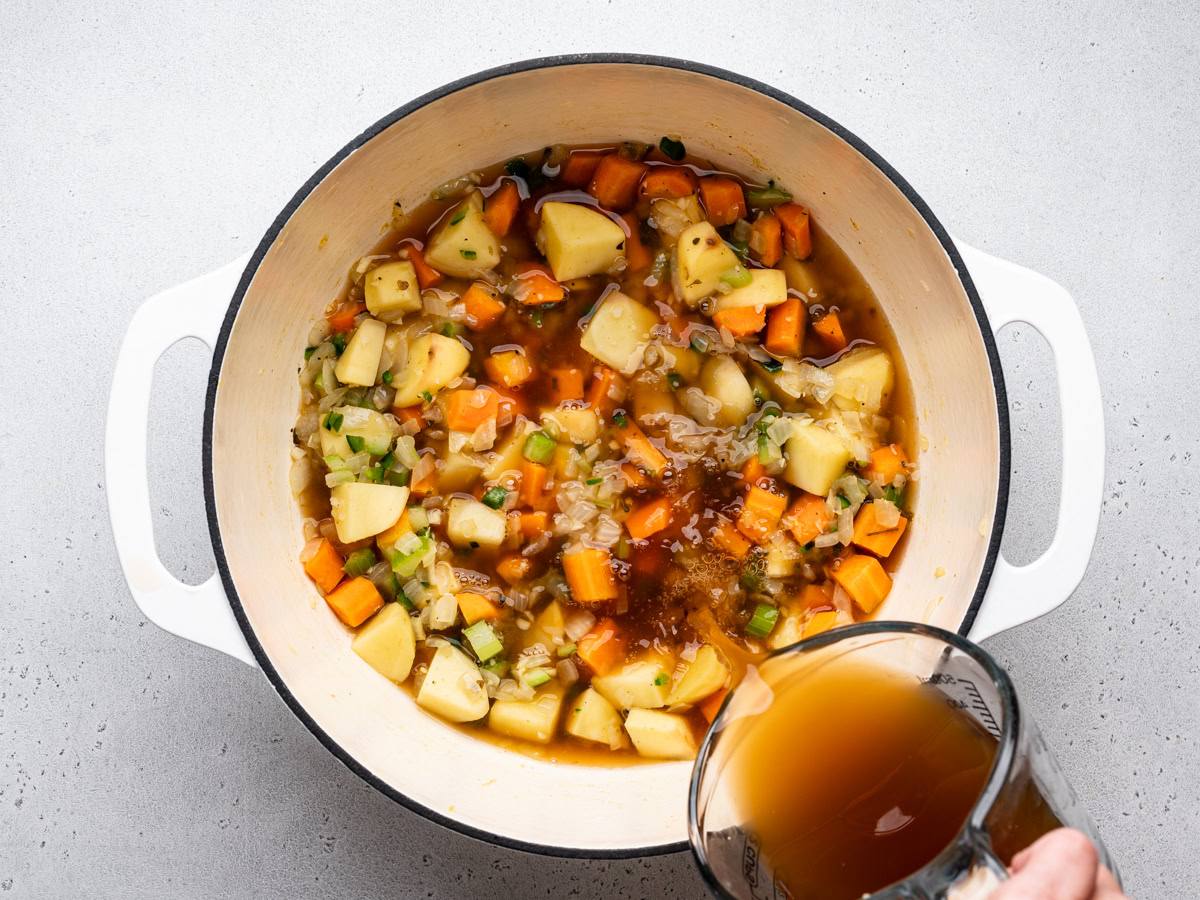 pouring broth over sautéed vegetables in white Dutch oven